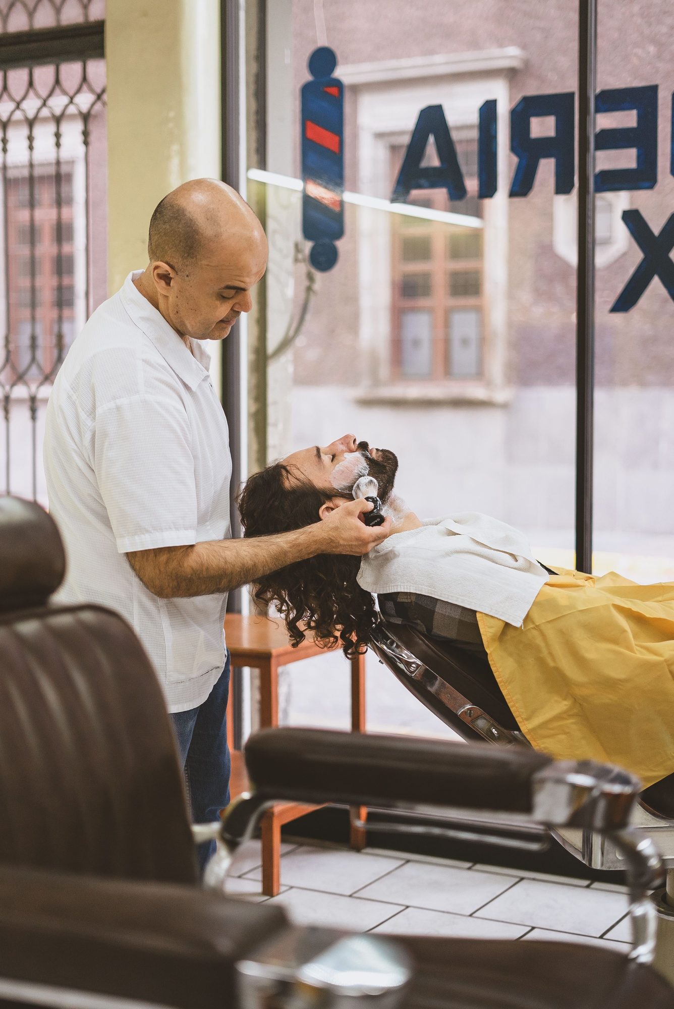 Barbeiro lavando o cabelo de um cliente na cadeira reclinada, dentro de uma barbearia com vitrine para a rua