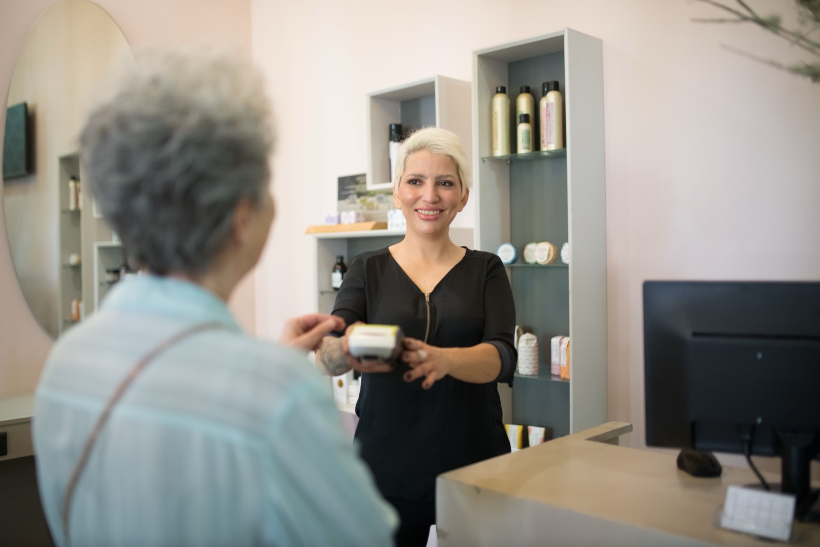 Recepcionista sorrindo segura a maquininha de cartão enquanto a cliente paga no balcão do salão