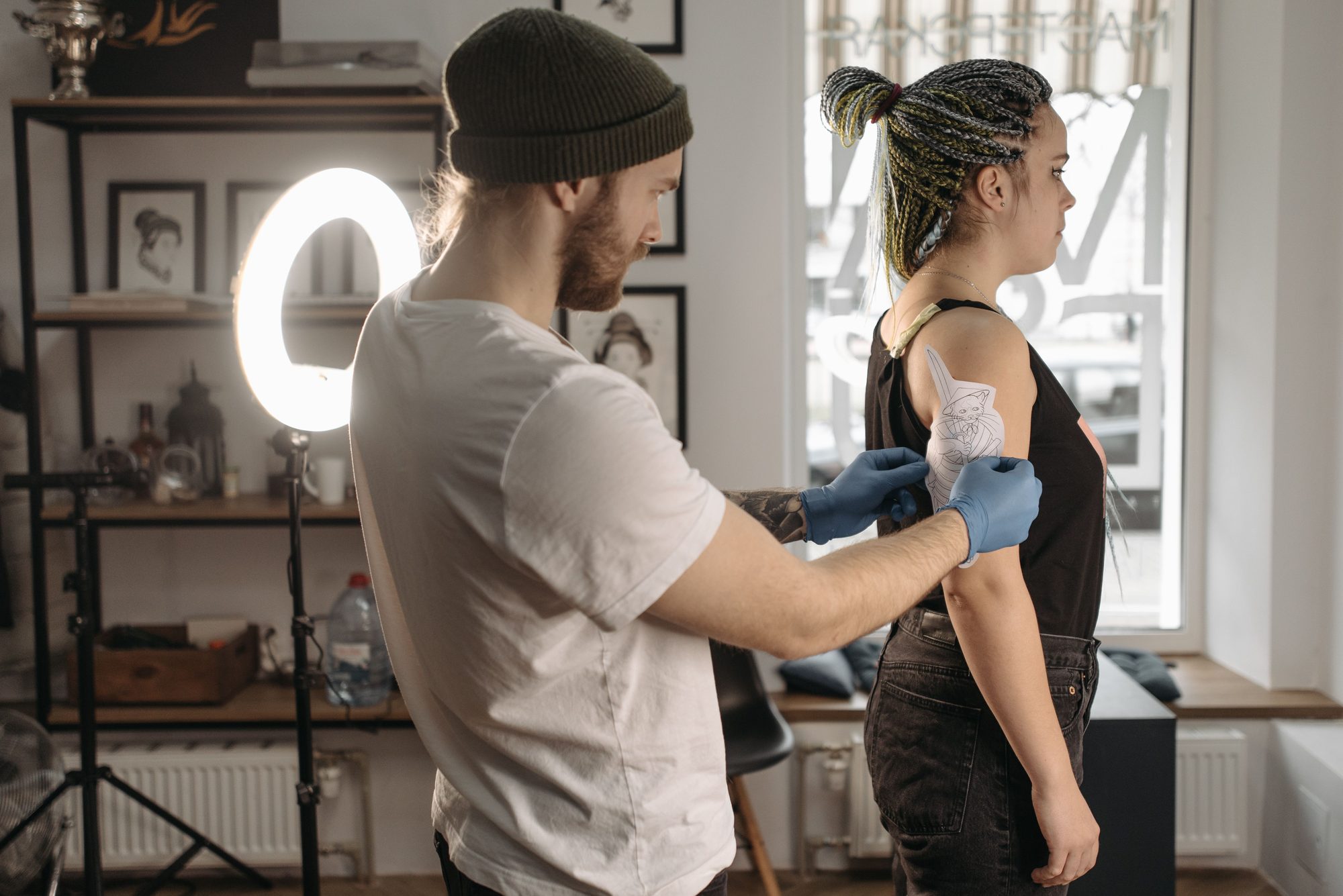 Tatuador de luvas preparando o braço de uma cliente em pé, em um estúdio com ring light e quadros na parede