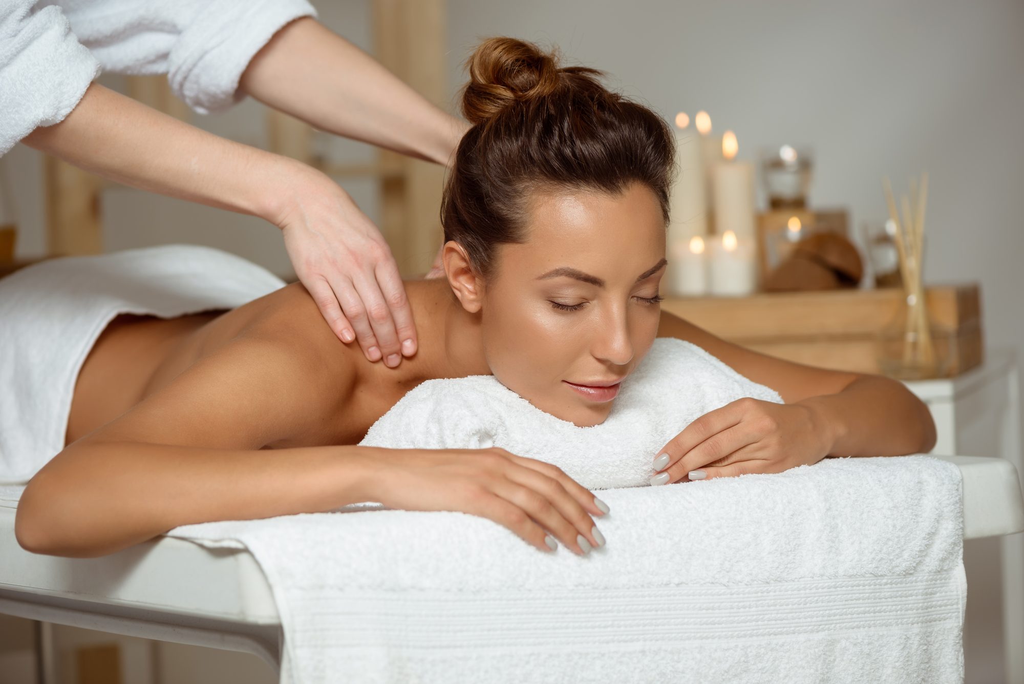 Cliente relaxando de olhos fechados durante uma massagem nas costas, em um spa com velas e toalhas brancas